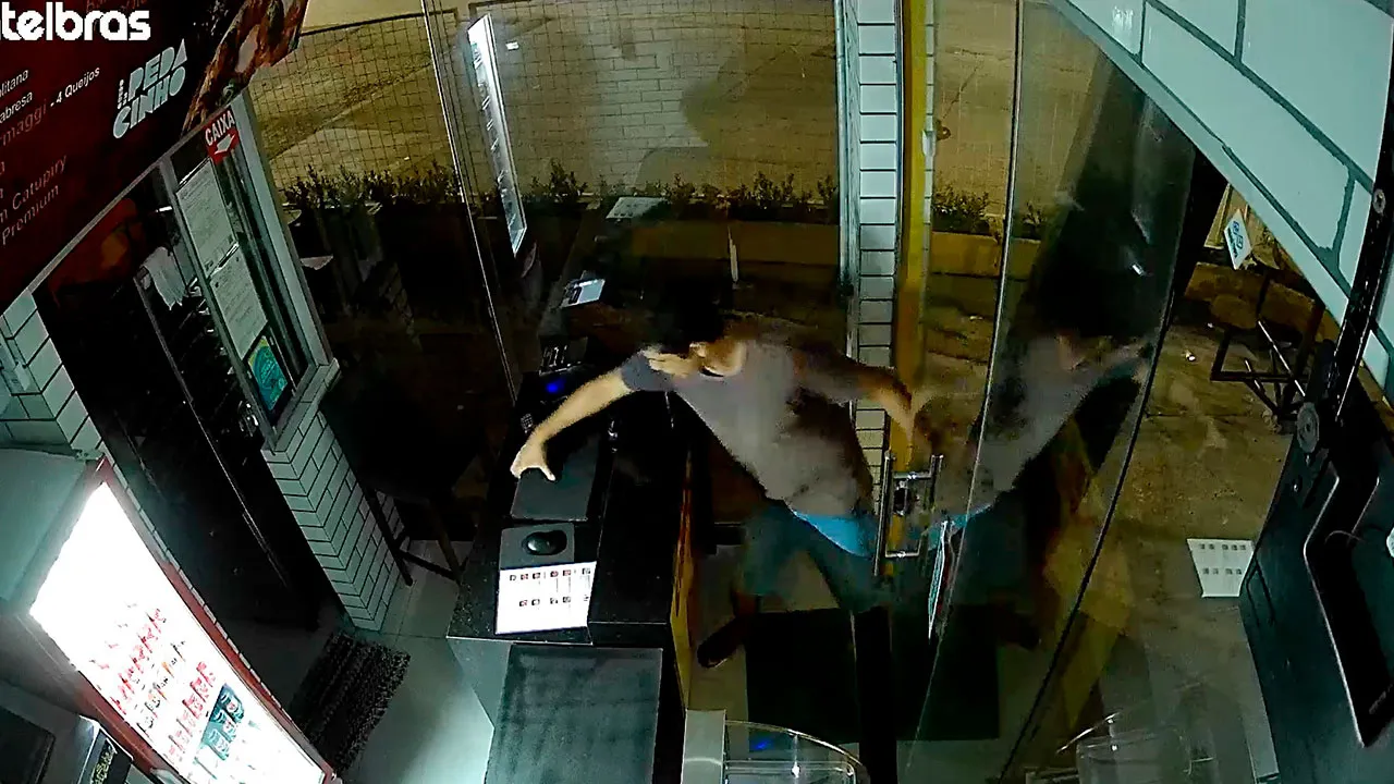Câmera de segurança flagra o momento exato em que o criminoso arromba a porta de vidro do estabelecimento.