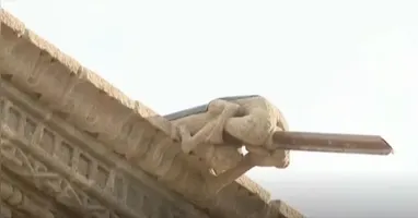 A peça em questão é uma escultura de pedra que representa um homem nu em posição agachada.