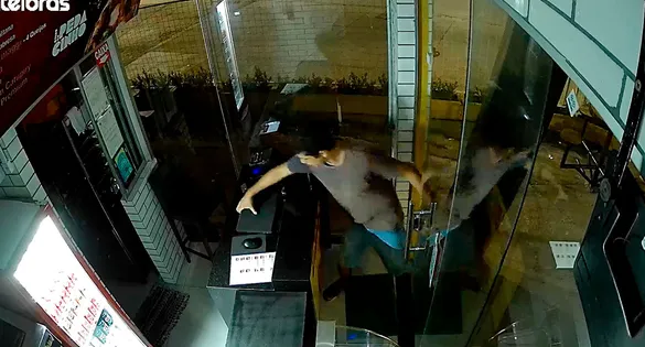 Câmera de segurança flagra o momento exato em que o criminoso arromba a porta de vidro do estabelecimento.